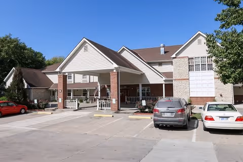 Exterior view of a senior living facility named Vintage Park At Atchison, showing the main entrance with a covered drop-off area supported by brick columns. Several cars are parked in front of the building, and there are trees and shrubs around the parking lot.