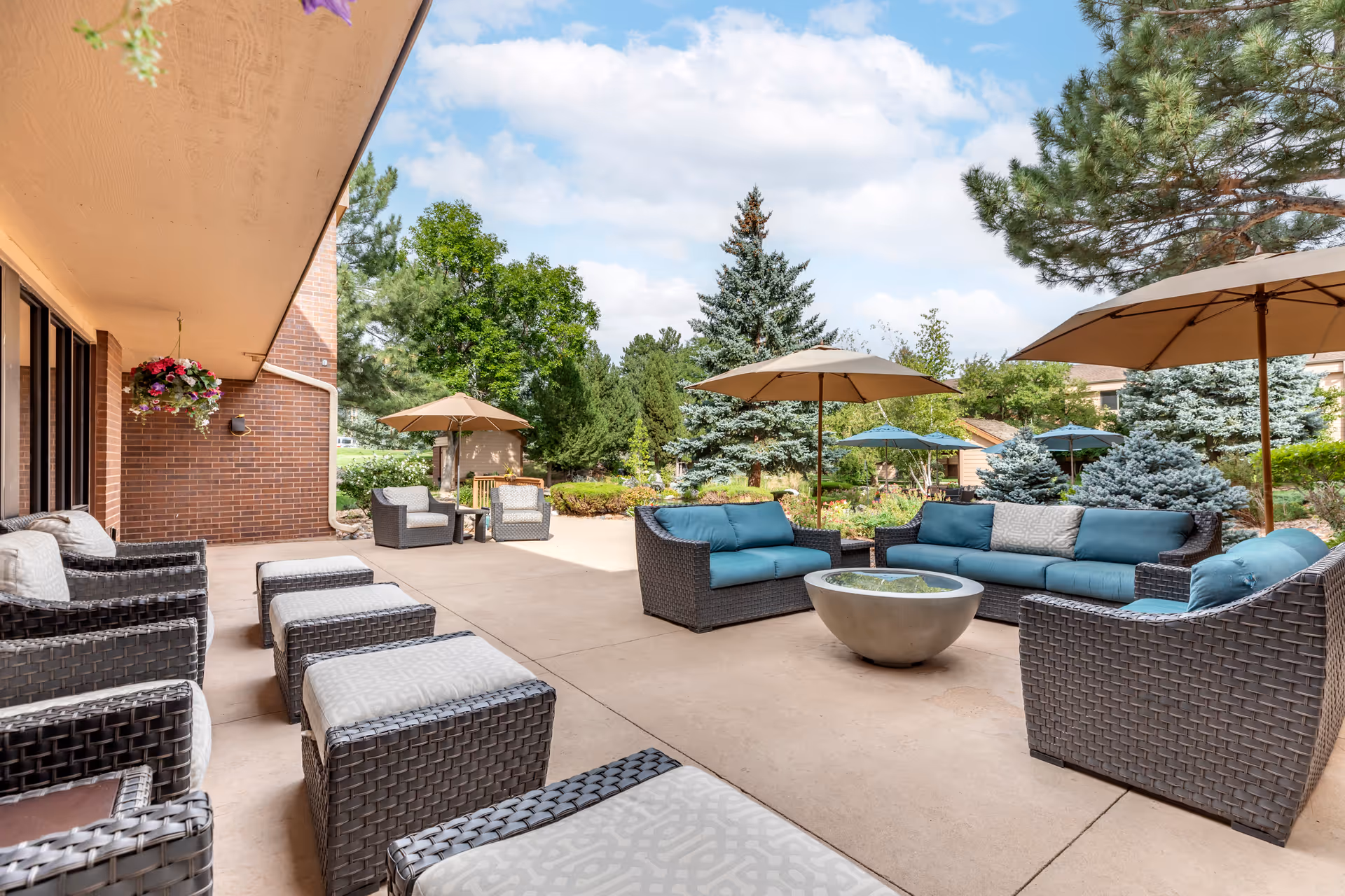 Outdoor patio area at Brookdale Meridian Boulder featuring wicker furniture with cushions, umbrellas, and a fire pit, surrounded by trees and greenery under a partly cloudy sky.