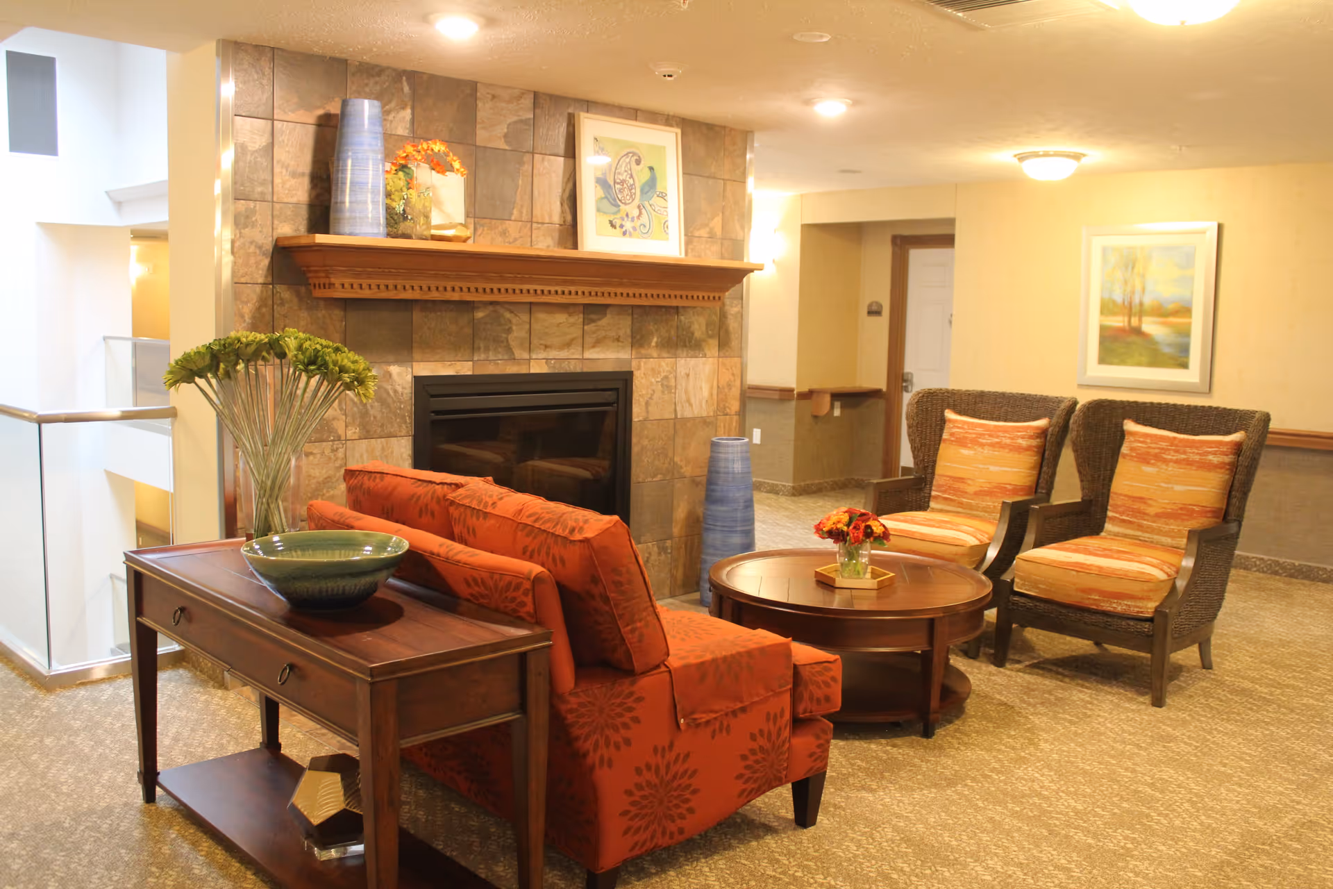A cozy seating area in a retirement community featuring a red patterned loveseat, two wicker armchairs with orange striped cushions, a round wooden coffee table with a small flower arrangement, and a wooden console table with decorative bowls and a vase of green flowers. The background includes a tiled fireplace with a wooden mantel displaying vases and framed artwork, beige walls with framed paintings, and soft lighting.