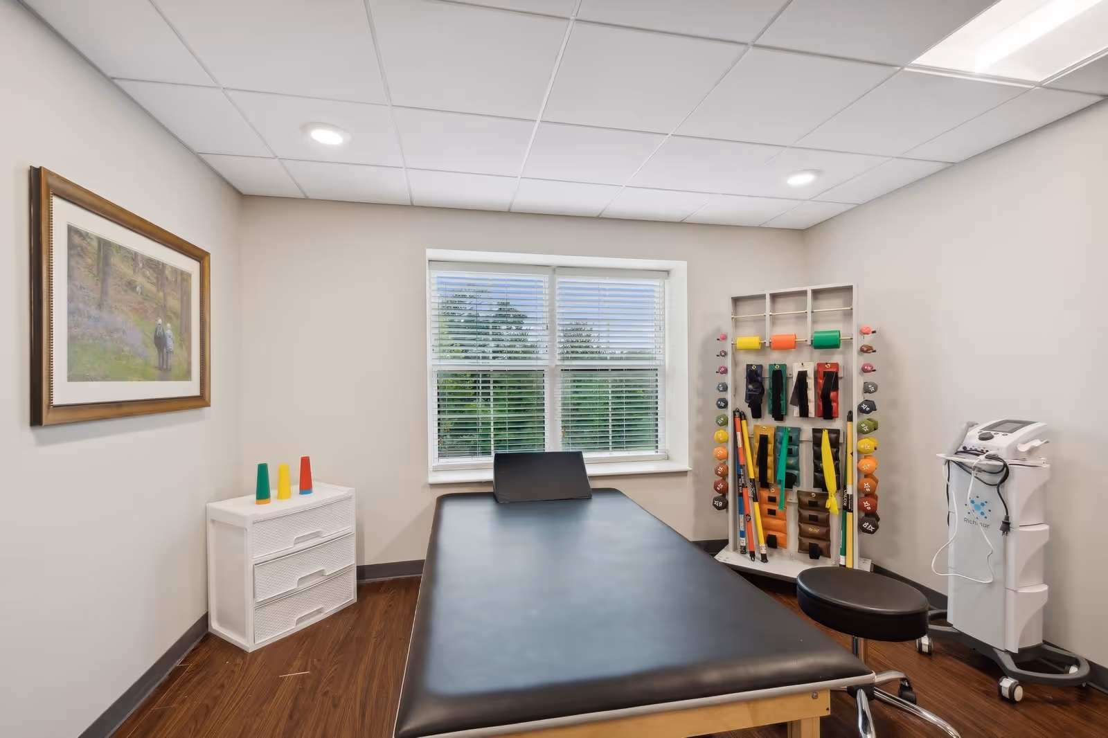 A therapy or exercise room with a black padded treatment table in the center, a small white cabinet with colorful cones on top, a rack holding various exercise bands and weights, a rolling stool, and a medical device on a cart. There is a window with blinds showing greenery outside and a framed picture on the wall.