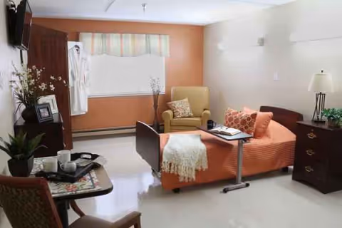 Orange bedspread on a hospital-style bed in a small room with chairs and dresser.