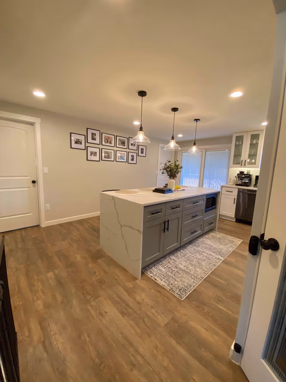 A modern kitchen with a large island in the center featuring gray cabinets and a white marble countertop. Three pendant lights hang above the island. The kitchen has wooden flooring, white walls, and a wall decorated with framed photos. There is a dishwasher and coffee machine visible in the background near glass-front cabinets.