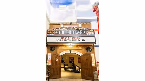 Entrance to an indoor theatre with a marquee reading 'THEATRE' and 'NOW SHOWING GONE WITH THE WIND', open wooden doors and seating visible inside.