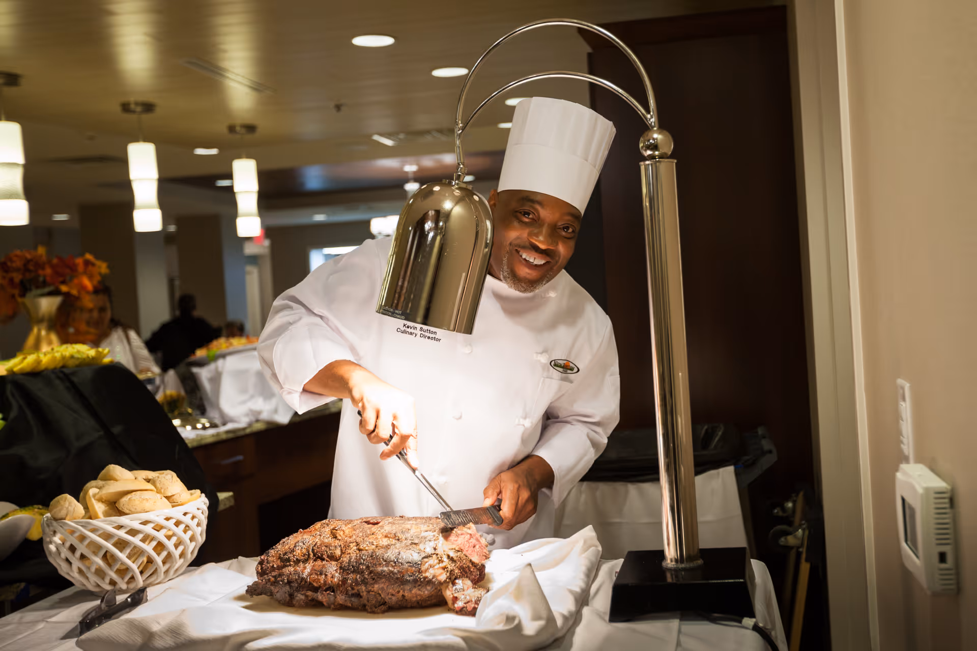 A chef wearing a white uniform and tall chef hat is carving a large piece of roasted meat under a heat lamp in a dining area. There is a basket of bread rolls on the table next to the meat, and the chef is smiling at the camera.