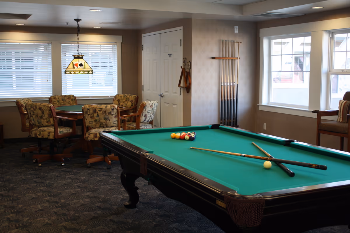 Interior recreation room with a pool table in the foreground and a card table with upholstered chairs by windows.