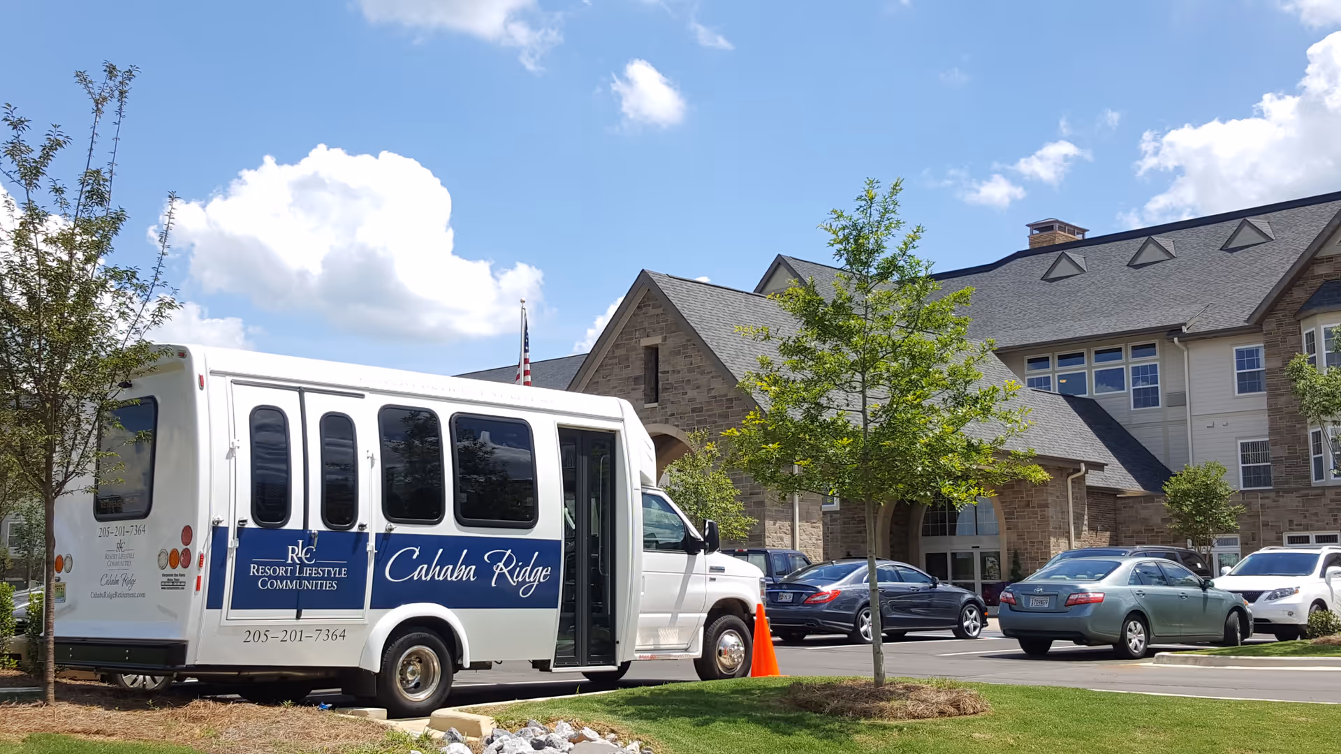 Exterior view of Cahaba Ridge Retirement Community building with a white shuttle bus parked in front. The bus has signage for Cahaba Ridge and Resort Lifestyle Communities. Several cars are parked nearby, and there are small trees and a clear blue sky with some clouds.