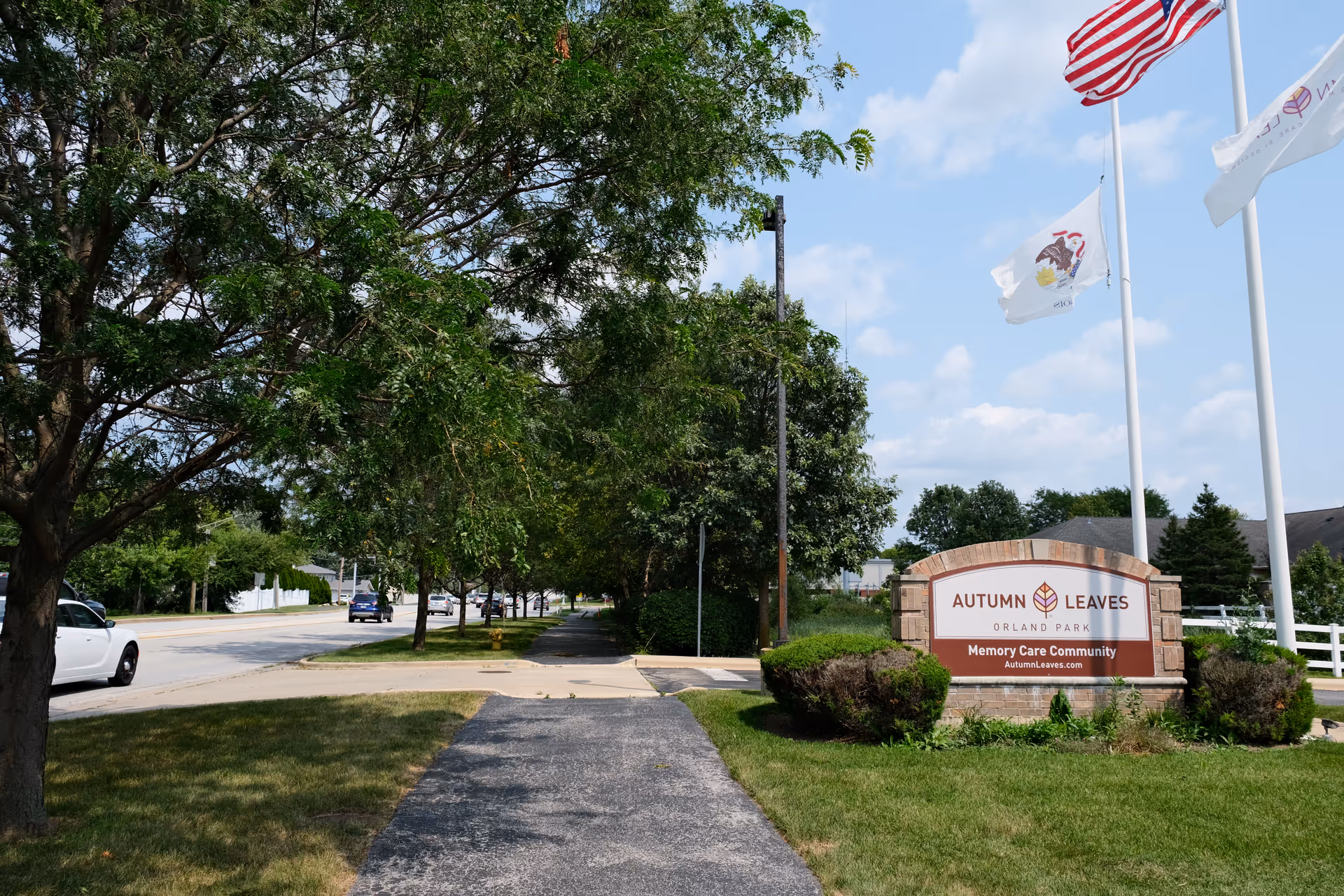 Sidewalk and street view with trees lining the sidewalk and a sign for Autumn Leaves Orland Park Memory Care Community on the right side, along with three flagpoles flying the American flag and other flags under a partly cloudy sky.