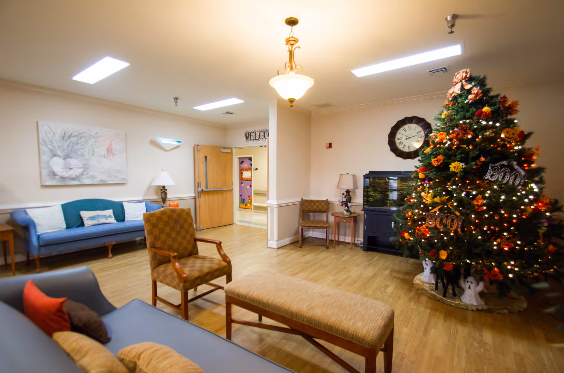 A cozy common area in a senior living facility decorated for Halloween with a Christmas tree adorned with orange and black ornaments, lights, and 'Boo' signs. The room has wooden flooring, a blue couch with pillows, a patterned armchair, a bench, a wall clock, a fish tank, and a 'Welcome' sign above a door.