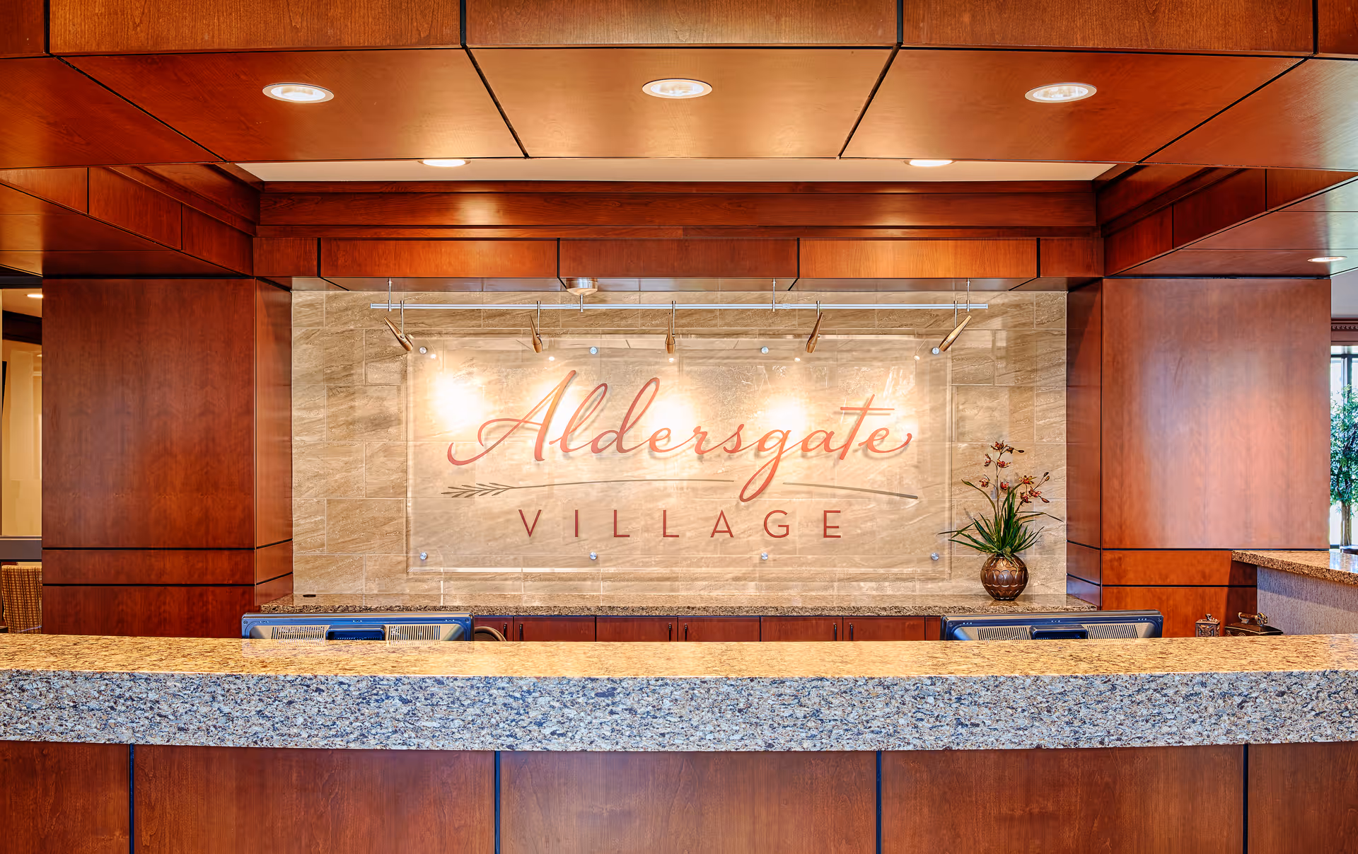 Reception desk area with a granite countertop and wooden paneling. Behind the desk is a wall with a large sign that reads 'Aldersgate Village' in elegant script. A small plant in a vase is placed on the right side of the desk.