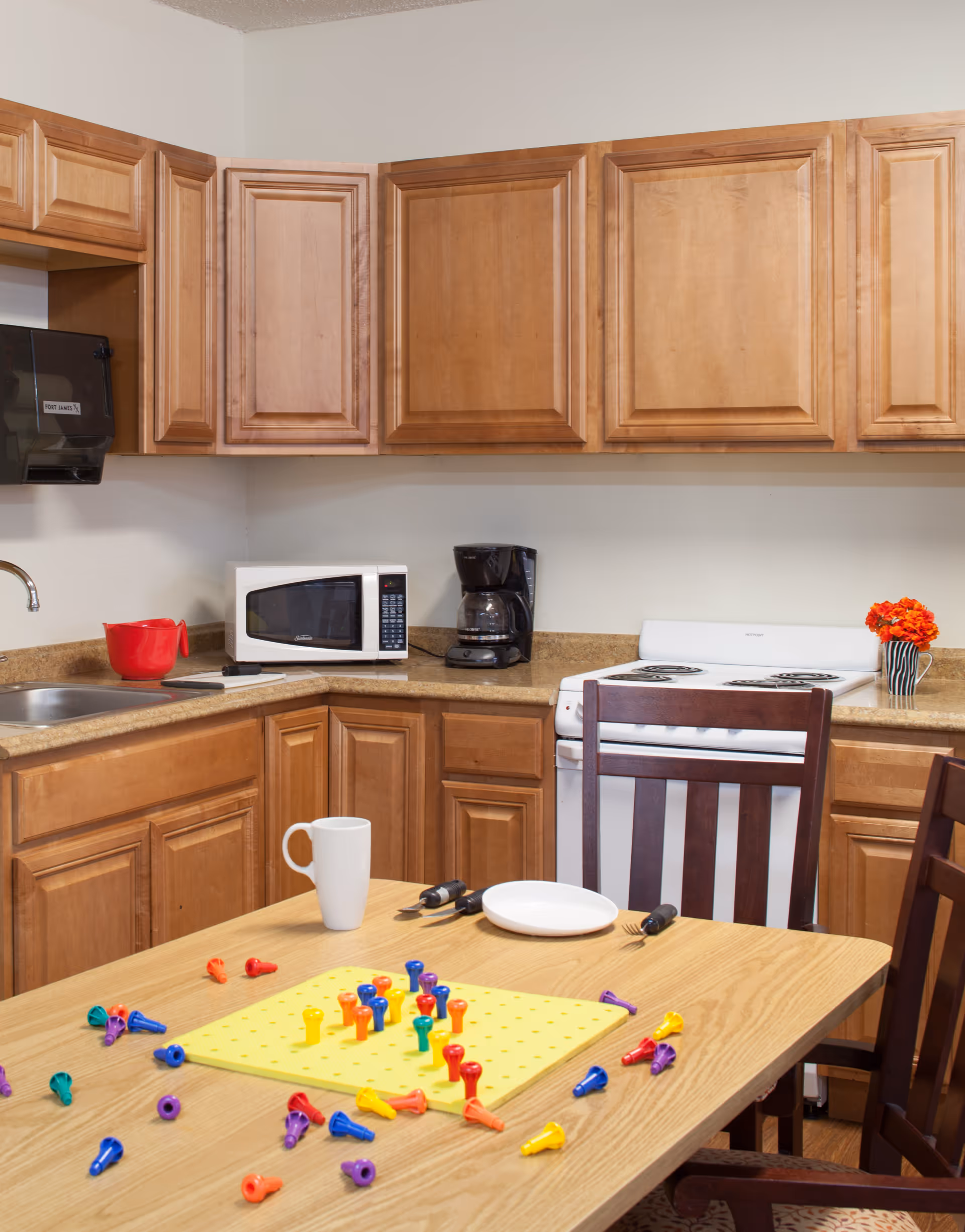 A kitchen with wooden cabinets, a microwave, coffee maker, stove, and sink. A wooden table in the foreground has a white mug, a plate, utensils, and a yellow pegboard game with colorful pegs scattered around.