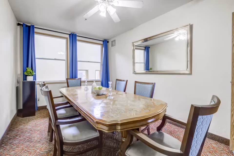 A dining room with a wooden table surrounded by six chairs with blue upholstery. The table has a bowl with green apples and two candle holders. There are two large windows with blue curtains and a large rectangular mirror on the wall reflecting the ceiling fan with lights.