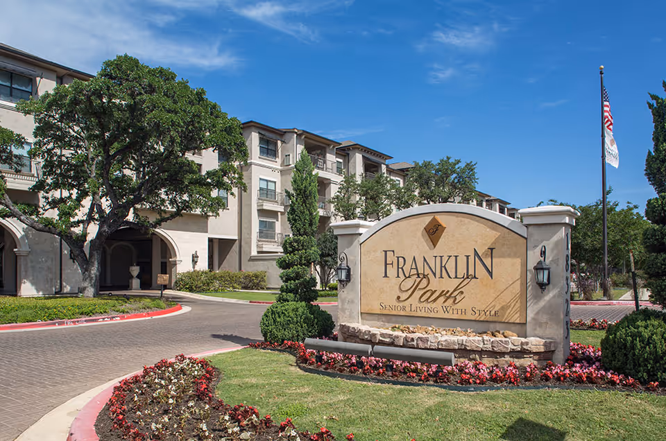 Exterior view of Franklin Park Sonterra senior living facility with a large stone sign in the foreground surrounded by flowers and greenery, and a multi-story building with balconies and trees in the background under a clear blue sky.