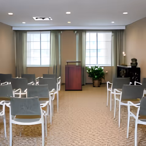Small meeting room with rows of chairs facing a wooden podium between two large windows and a potted plant.