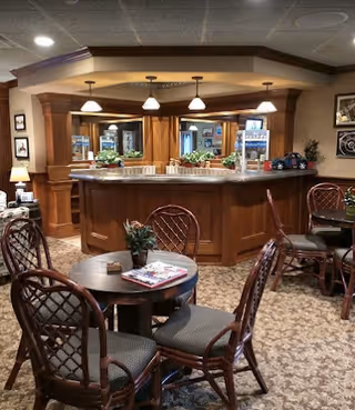 Cozy interior space with a wooden bar counter, three hanging pendant lights, and several round tables with cushioned chairs arranged around the room. The area has carpeted flooring and decorative plants on the tables and counter.