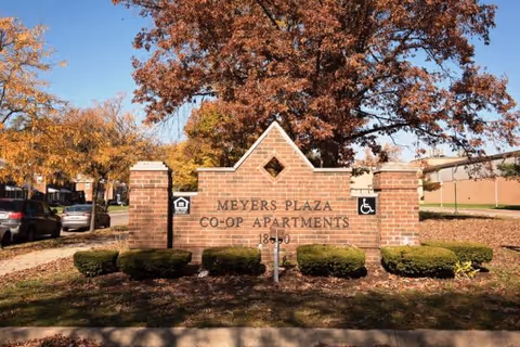 Brick entrance sign that reads "Meyers Plaza Co-Op Apartments" set among landscaped shrubs and autumn trees with parked cars along the street.