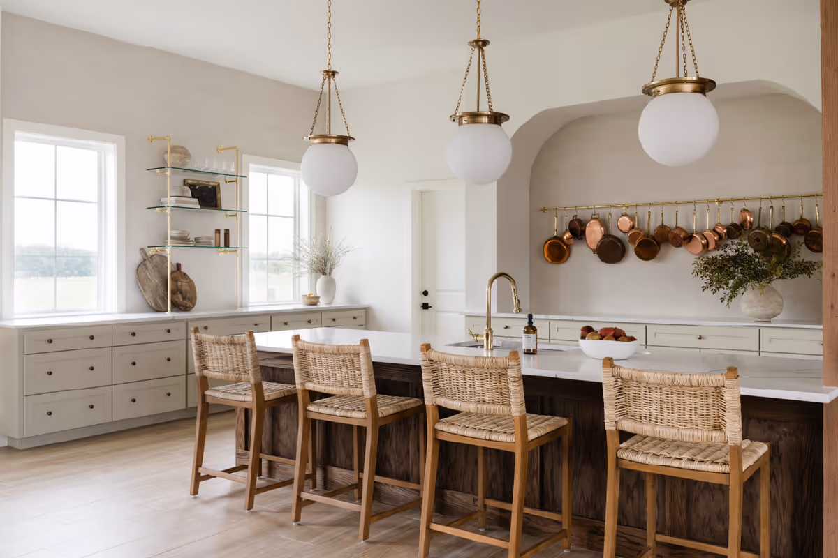 Bright and spacious kitchen with a large island featuring a white countertop and four wicker bar stools. Three round pendant lights hang above the island. The kitchen has light-colored cabinetry, two large windows, open glass shelves with dishes, and a wall-mounted rack holding copper pots and pans. A vase with greenery and a bowl of fruit are placed on the countertops.