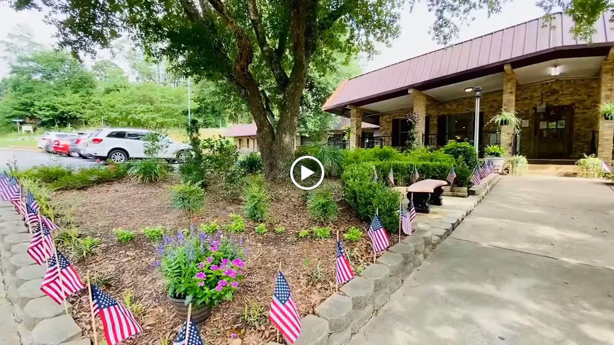 Exterior view of Henderson Health & Rehab Center showing a garden area with small American flags planted along the edge, a large tree, and a brick building entrance with a covered walkway. Several cars are parked in the background.