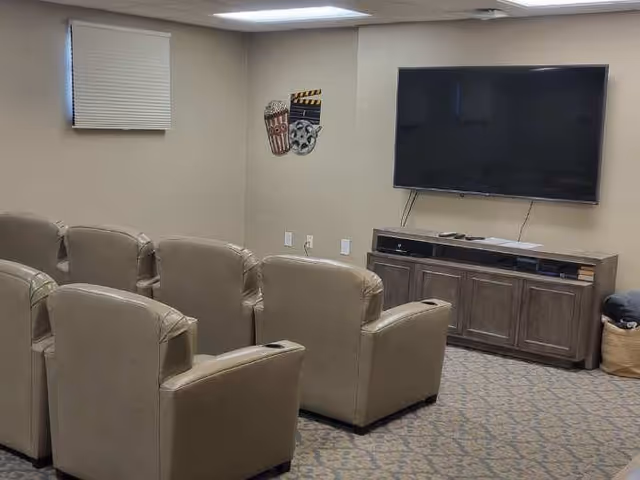 A small media room with two rows of beige leather recliner chairs facing a large flat-screen TV mounted on a beige wall. Below the TV is a wooden cabinet with electronic devices and books. The room has a patterned carpet, a small window with a closed blind, and a wall decoration featuring popcorn and film reel icons.