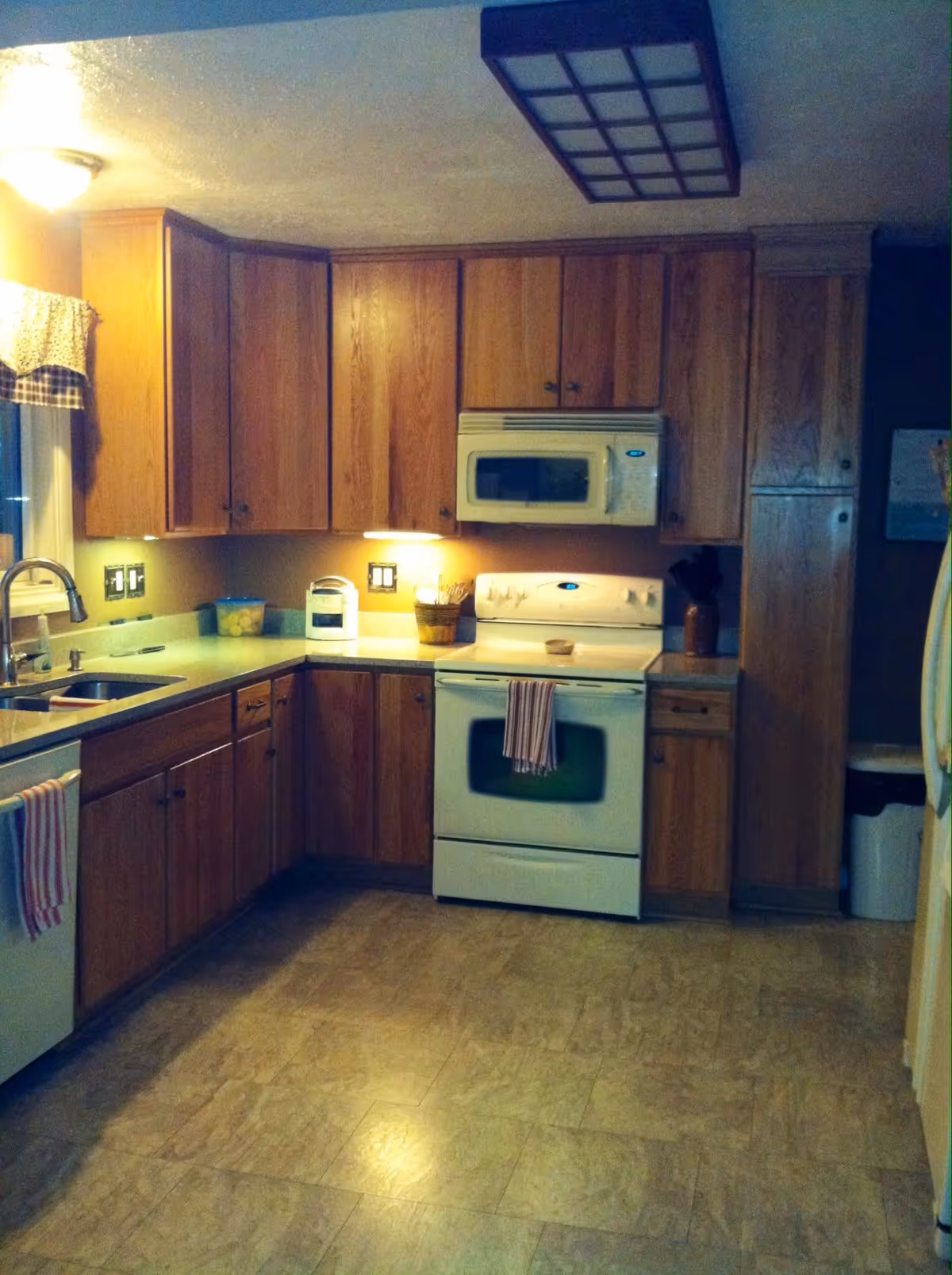 A kitchen with wooden cabinets, a white stove with a microwave above it, a sink under a window with a checkered valance, and a countertop with various items including a container of lemons and a small appliance. The floor is tiled, and there are two striped towels hanging on the stove and dishwasher handles.