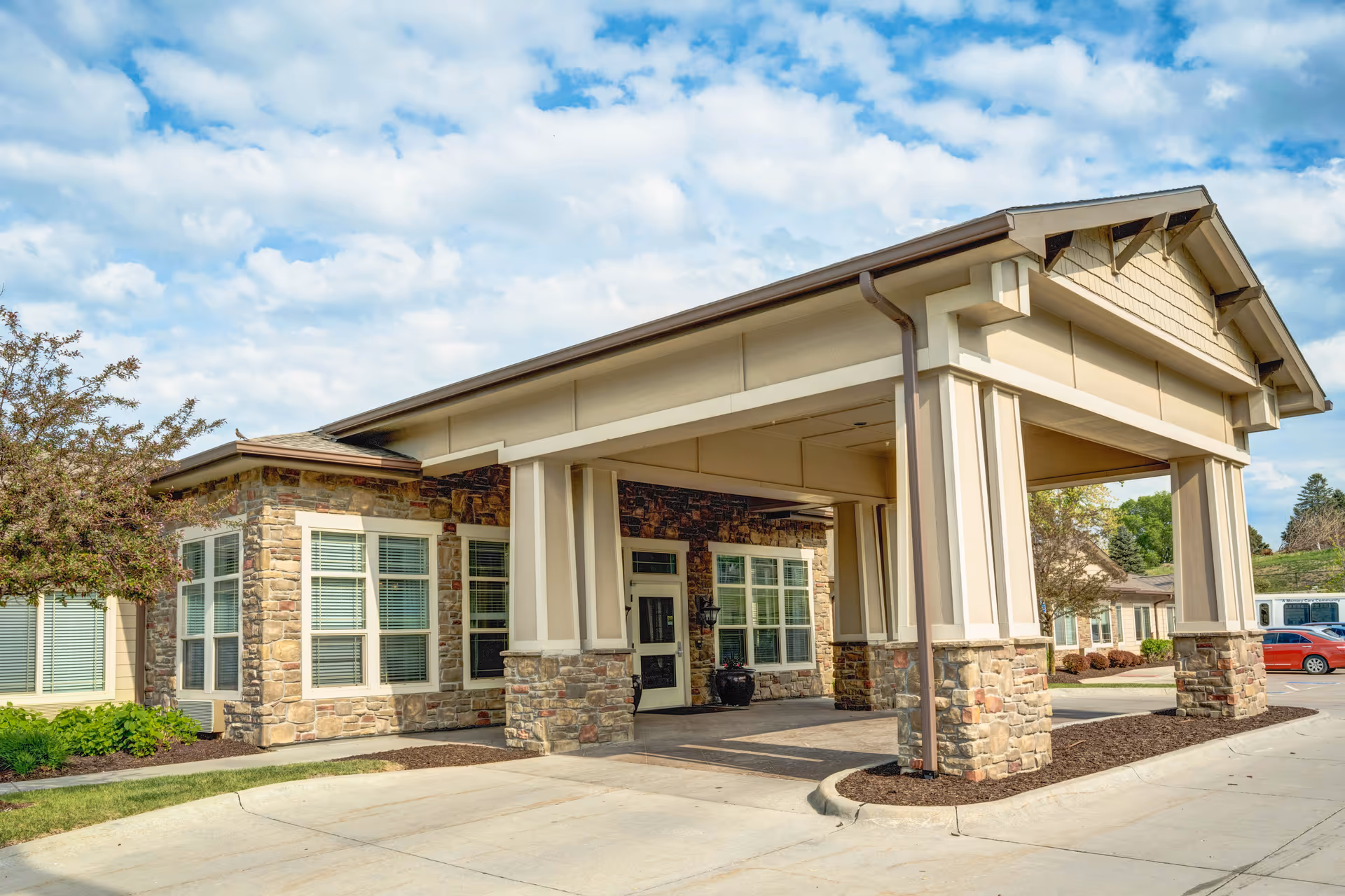 Exterior view of Cedar Creek of Prairie Meadows facility entrance with a covered drop-off area supported by stone and beige pillars, surrounded by landscaping and a partly cloudy sky.