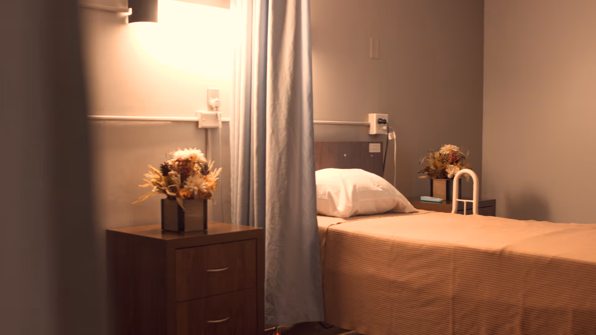 A simple bedroom in a senior living facility with a single bed covered in a beige blanket, a white pillow, a wooden nightstand with a floral arrangement, and a privacy curtain partially drawn.