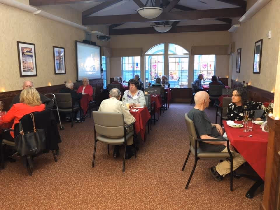 A dining room in a senior living facility with several elderly people seated at tables covered with red tablecloths, engaged in conversation and dining. The room has beige walls, framed pictures, large windows with decorative glass, and ceiling lights with exposed wooden beams.