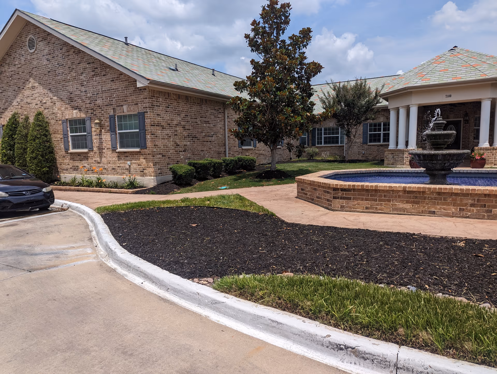 Exterior view of a brick building with a sloped roof, several windows with shutters, and a covered entrance supported by white columns. In front of the entrance is a tiered water fountain surrounded by a brick base. The area includes landscaped grass, mulch beds, small trees, and shrubs. A black car is partially visible parked on the left side near a curved concrete driveway.