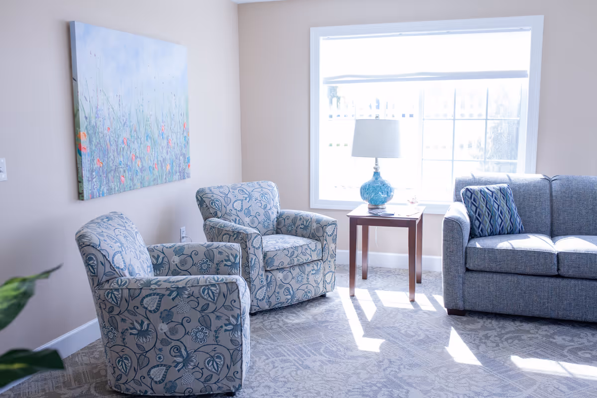 A bright living room with two patterned armchairs, a gray sofa with a decorative pillow, a wooden side table with a blue glass lamp, and a large window letting in natural light. A colorful floral painting hangs on the beige wall.
