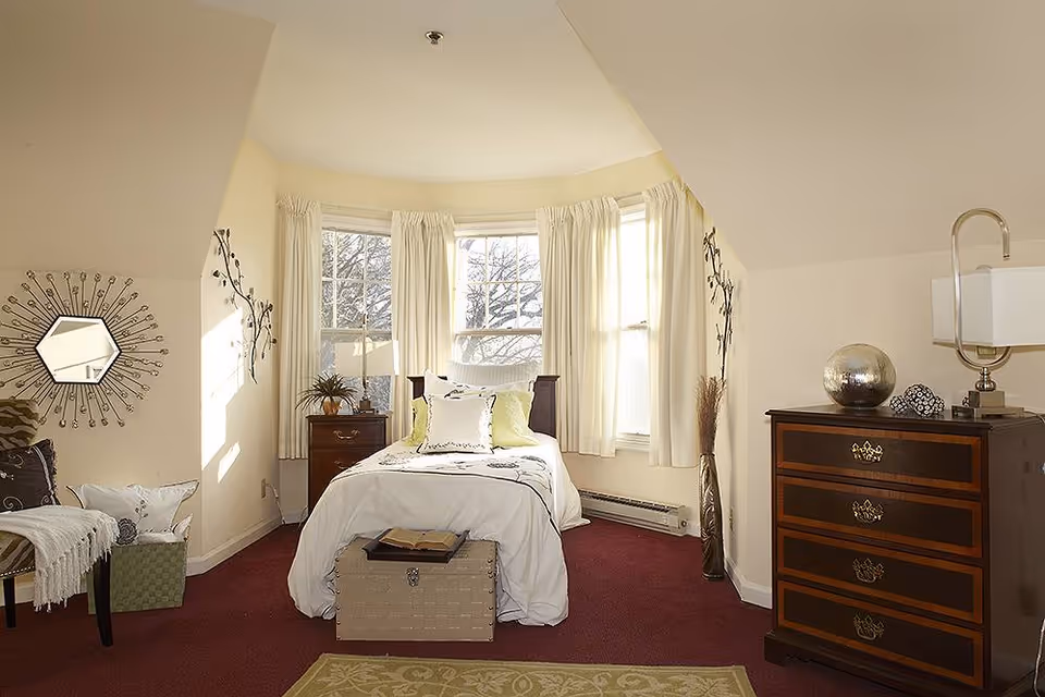 A cozy bedroom with a single bed centered under three large windows with white curtains. The bed is made with white bedding and decorative pillows. To the left of the bed is a wooden nightstand with a lamp and a plant. On the right side, there is a wooden dresser with decorative items and a lamp. The room has beige walls, a red carpet, and a green rug in the foreground. A decorative mirror and wall art are visible on the left wall.