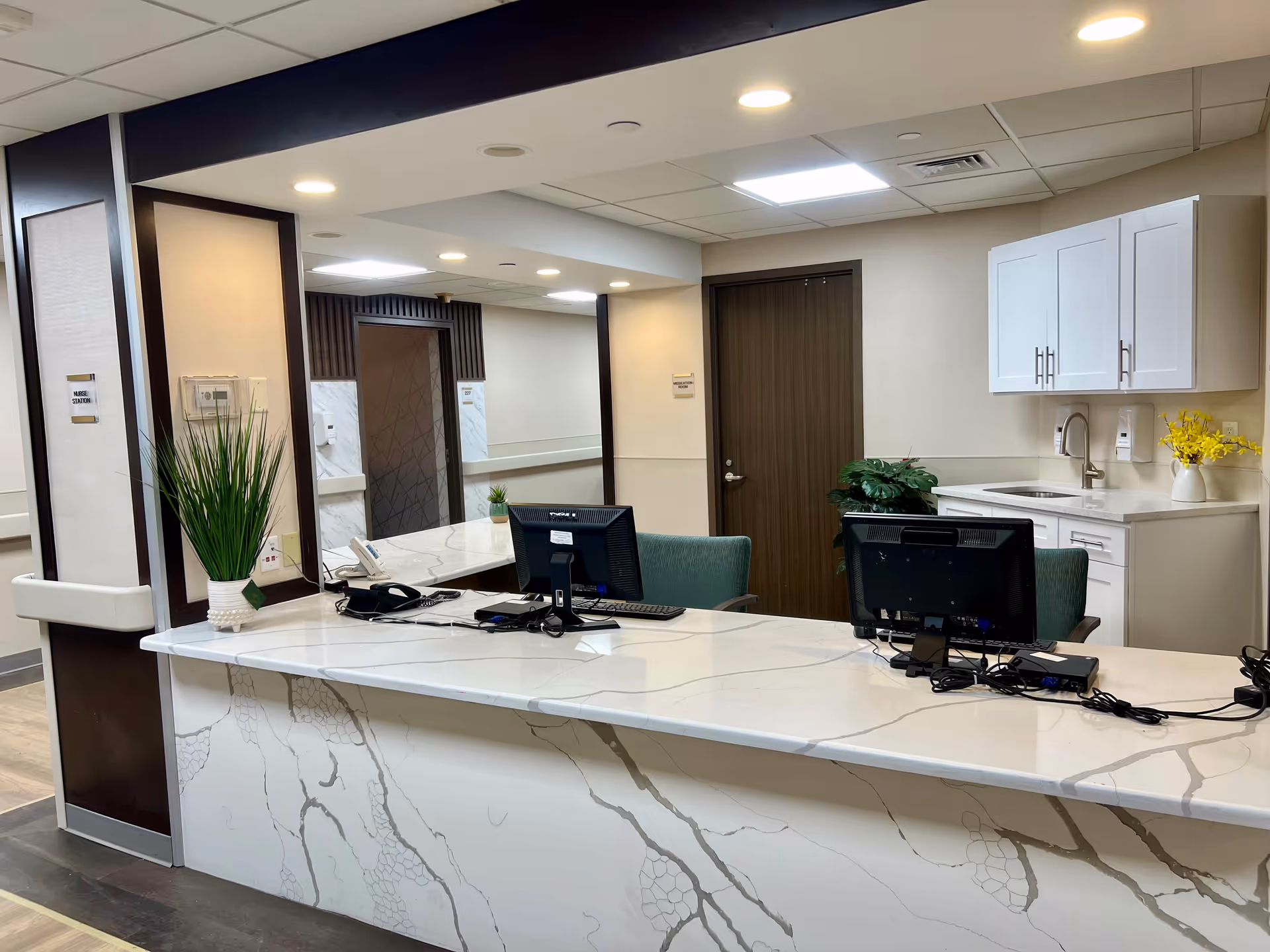 Reception desk area in a senior living facility with two computer monitors, a telephone, and office supplies on a marble countertop. Behind the desk are two green chairs, a door, white cabinets, a sink, and decorative plants. The walls are light-colored with dark trim and recessed lighting in the ceiling.