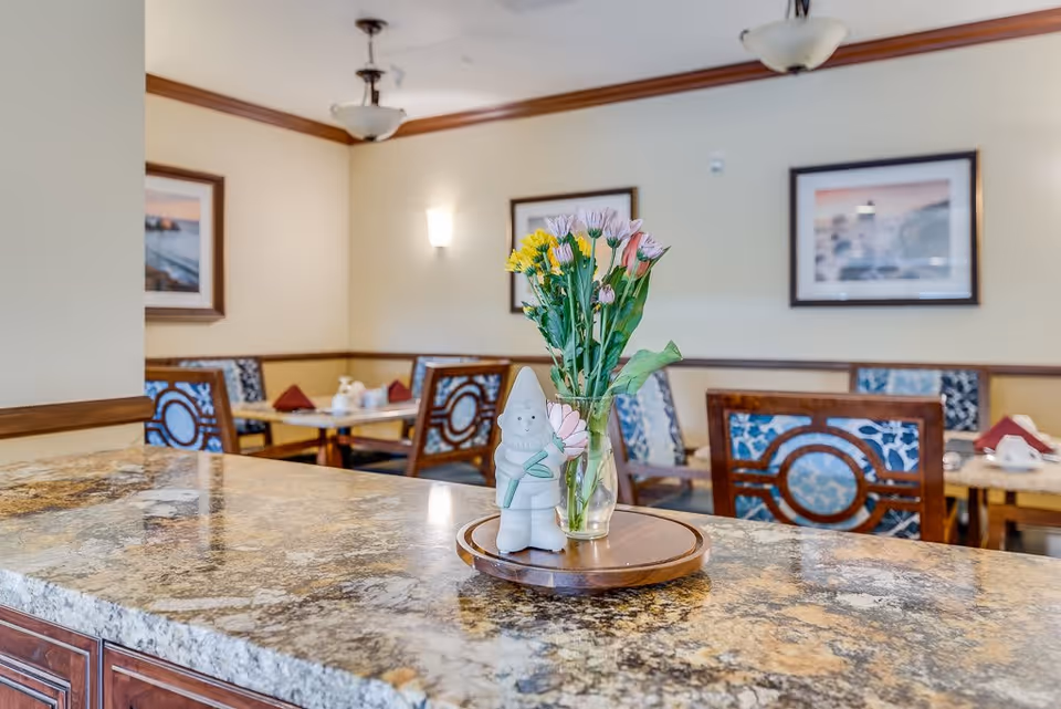 A dining area in a senior living facility with a granite countertop in the foreground holding a small vase of flowers and a ceramic gnome figurine. In the background, there are tables and chairs with patterned upholstery, framed artwork on the walls, and soft lighting fixtures.
