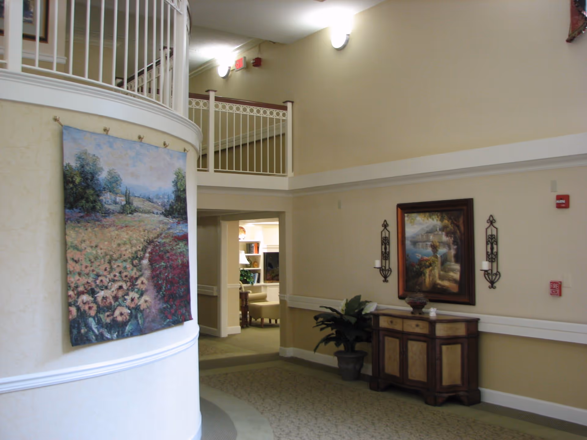 Interior hallway of a senior living facility with beige walls and carpeted floor. A large tapestry depicting a field of flowers hangs on a curved wall to the left. On the right wall, there is a wooden cabinet with decorative items on top, flanked by two candle holders and a framed painting of a scenic landscape. A potted plant is placed beside the cabinet. The hallway leads to a room with a fireplace and bookshelves visible in the background. White railings and a second-floor balcony are visible above.