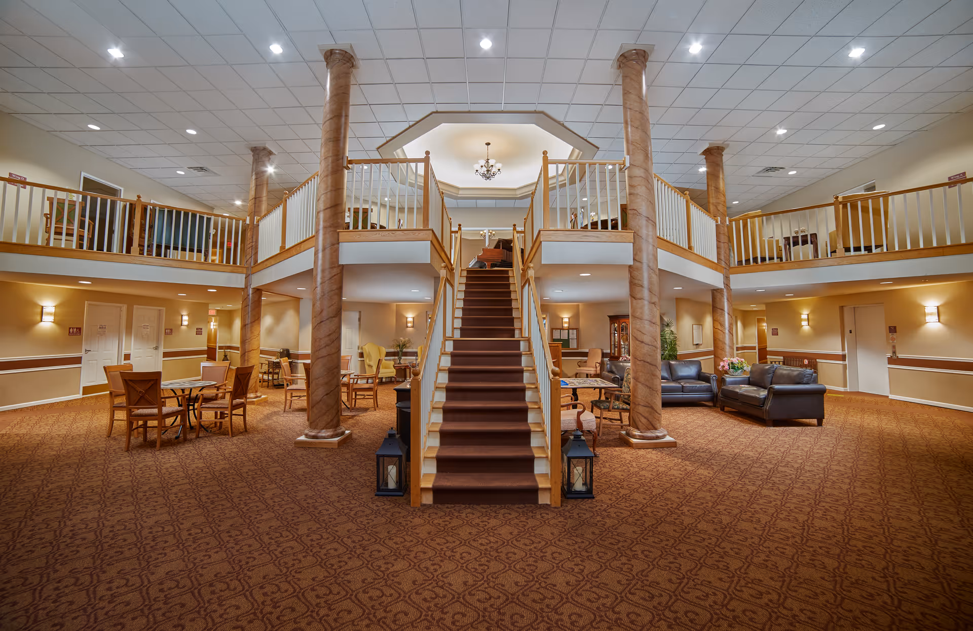 Spacious senior living facility common area with a central staircase leading to an upper balcony. The room features patterned carpet, multiple seating areas with chairs and sofas, tall decorative columns, warm lighting, and a chandelier above the staircase.