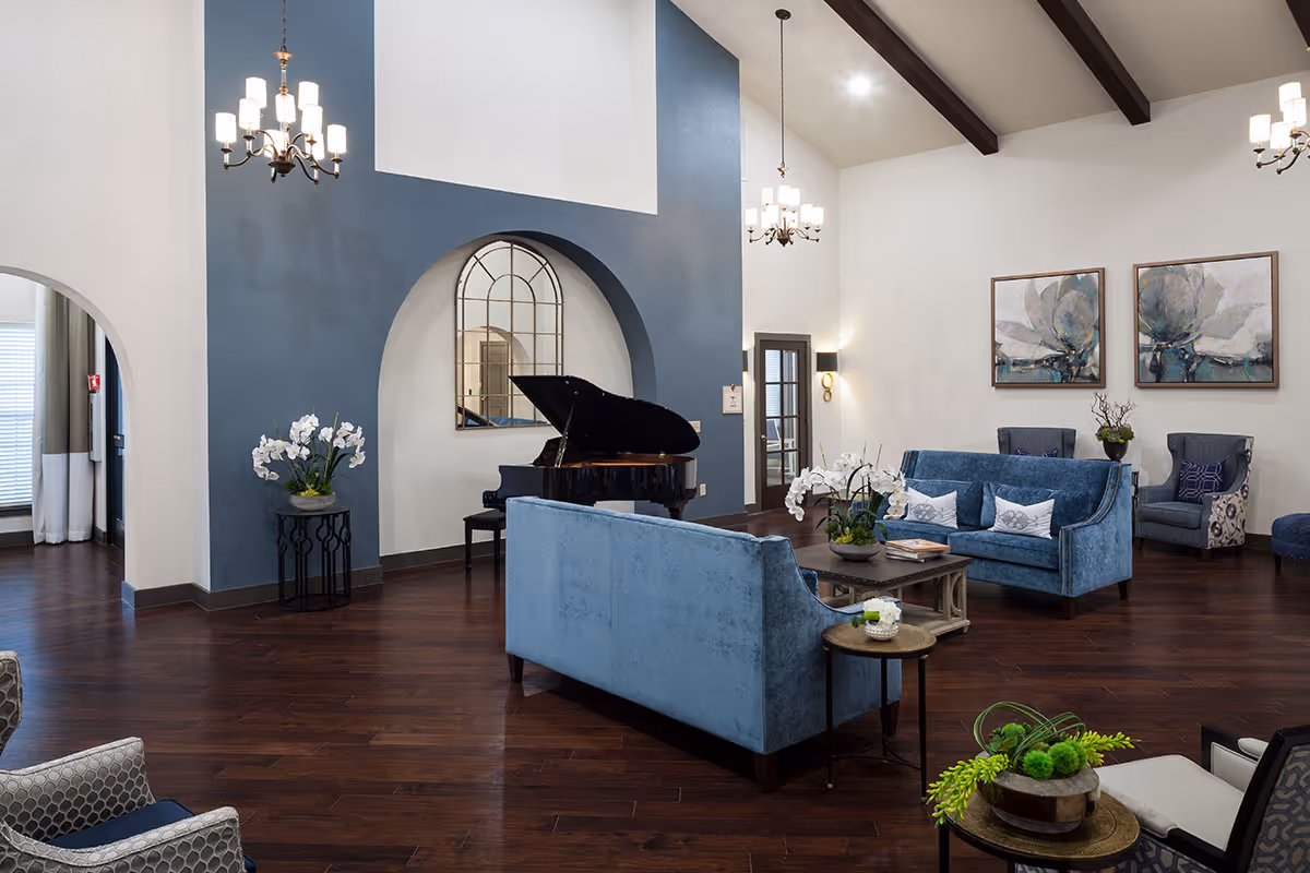 Spacious common lounge with blue sofas, a grand piano, chandeliers, and hardwood floors.