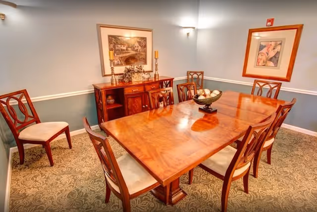 A formal dining room with a polished wooden table surrounded by eight wooden chairs with cushioned seats. The room has light blue walls with white trim and patterned carpet. A wooden sideboard with decorative items and framed artwork is against the wall.