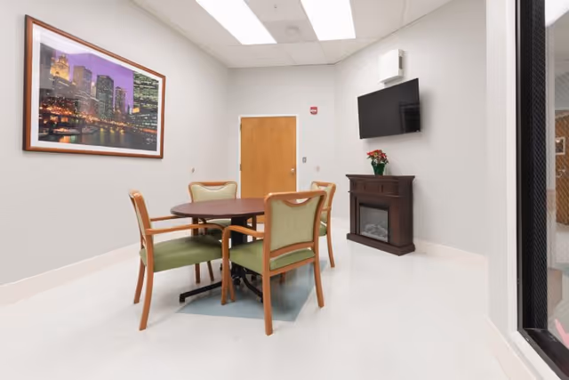 Small lounge with a round table and four green-upholstered chairs, a wall-mounted TV above a faux fireplace, framed cityscape art, and a wooden door.