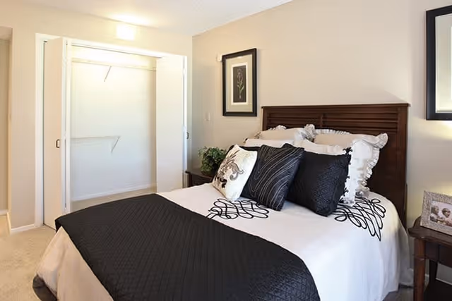 A neatly made bed with a dark wooden headboard in a bedroom. The bed has white and black bedding with multiple decorative pillows. To the left of the bed is an open closet with white sliding doors. There is a framed picture on the wall above a small bedside table with a plant. Another small table with a framed photo is on the right side of the bed.