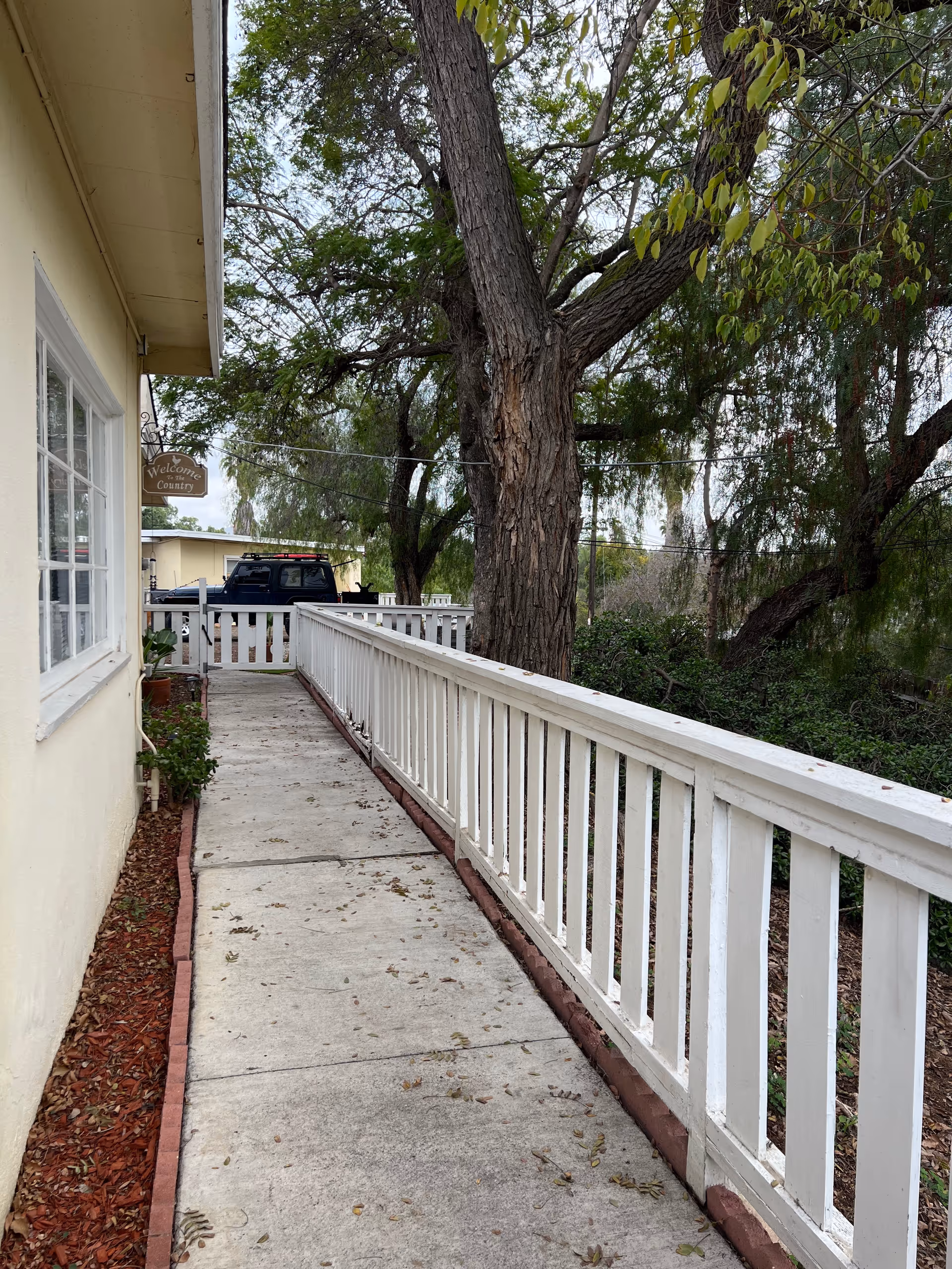 A concrete walkway with a white wooden railing on the right side and a yellow building wall with windows on the left side. There are trees and greenery on the right side beyond the railing, and a black truck is parked in the background near a white picket fence. A small wooden sign hanging from the building reads 'Welcome Country'.