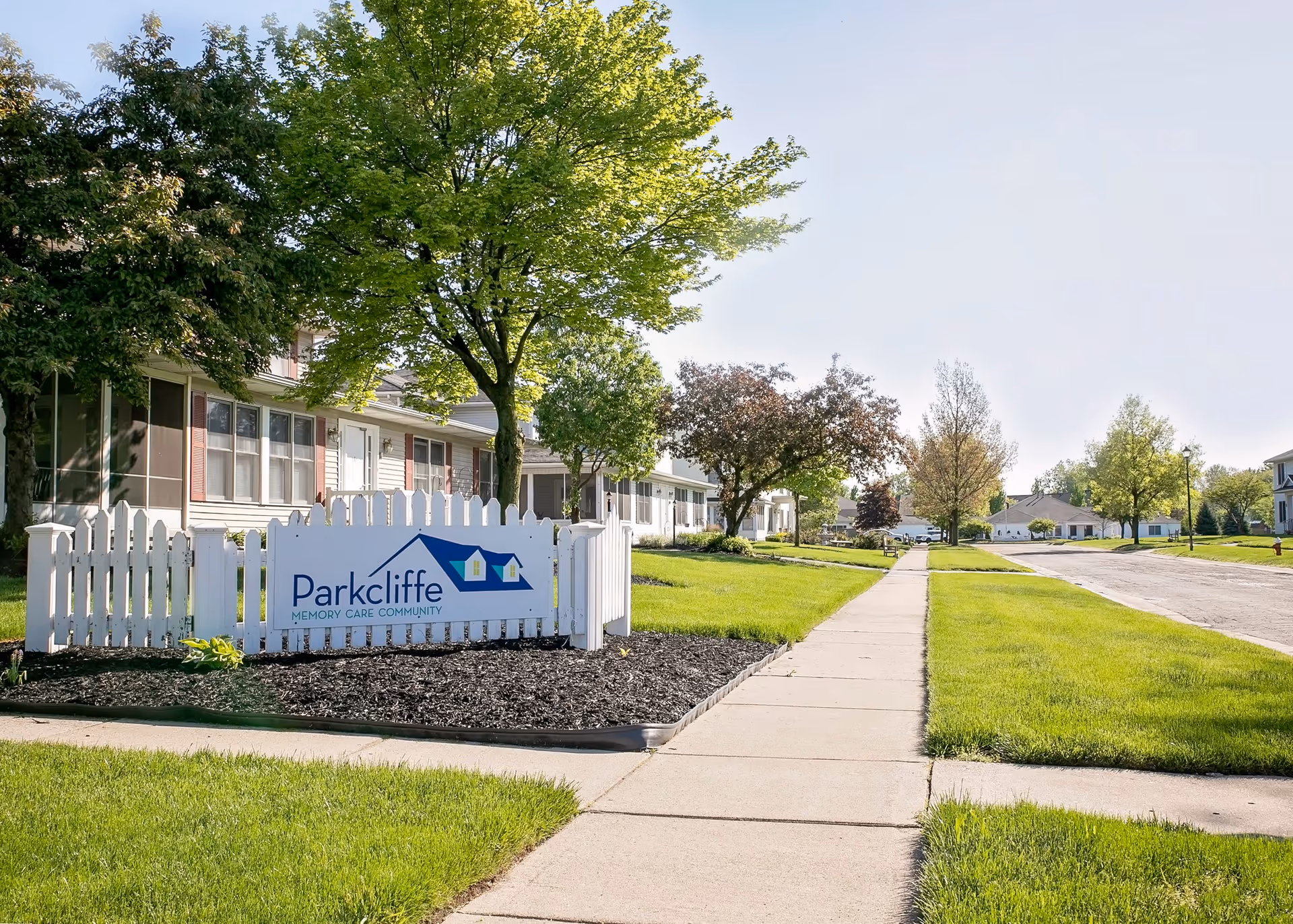 A sunny suburban street view of Parkcliffe Memory Care Community at Northwood, featuring a white picket fence with the community's sign, green lawns, trees, and single-story residential buildings along the sidewalk.