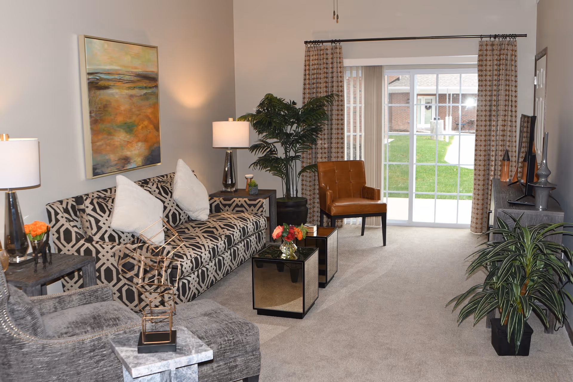Stylish furnished living room with a patterned sofa, armchairs, coffee tables, plants, and sliding glass doors to the outside.