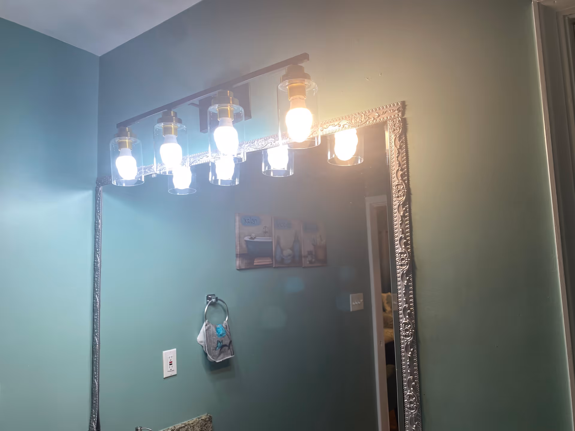 A bathroom interior with a large ornate framed mirror reflecting a wall with three decorative pictures. Above the mirror, there is a light fixture with six exposed bulbs enclosed in glass covers. A towel ring with a hanging towel is mounted on the green wall below the pictures.