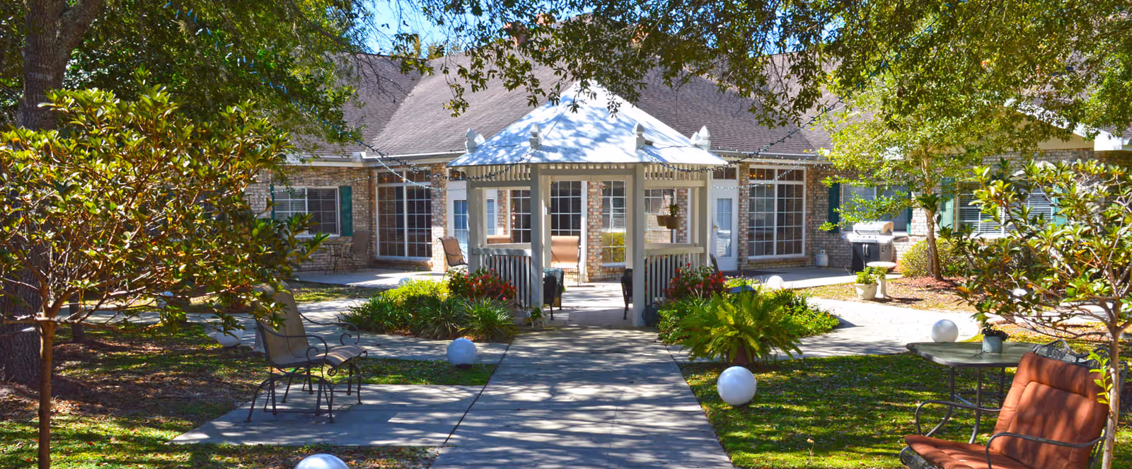 Outdoor courtyard area at Brookdale Canopy Oaks featuring a white gazebo in the center, surrounded by green trees, shrubs, and outdoor seating including chairs and tables. The building with large windows and a sloped roof is visible in the background.