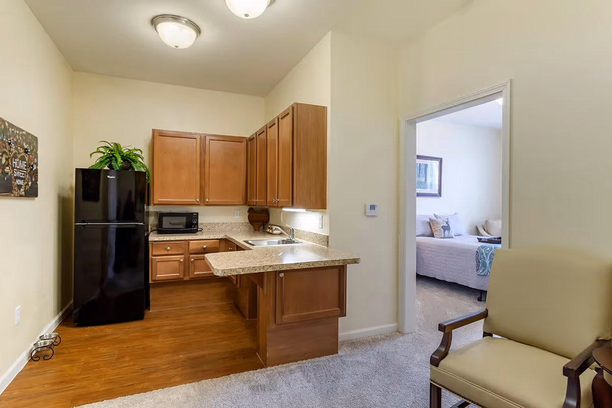 Interior view of a small kitchen area with wooden cabinets, a black refrigerator, a microwave on the counter, and a sink. There is a plant on top of the refrigerator and a decorative sign on the wall. Adjacent to the kitchen is a carpeted living space with a beige armchair and a doorway leading to a bedroom with a bed and framed artwork on the wall.
