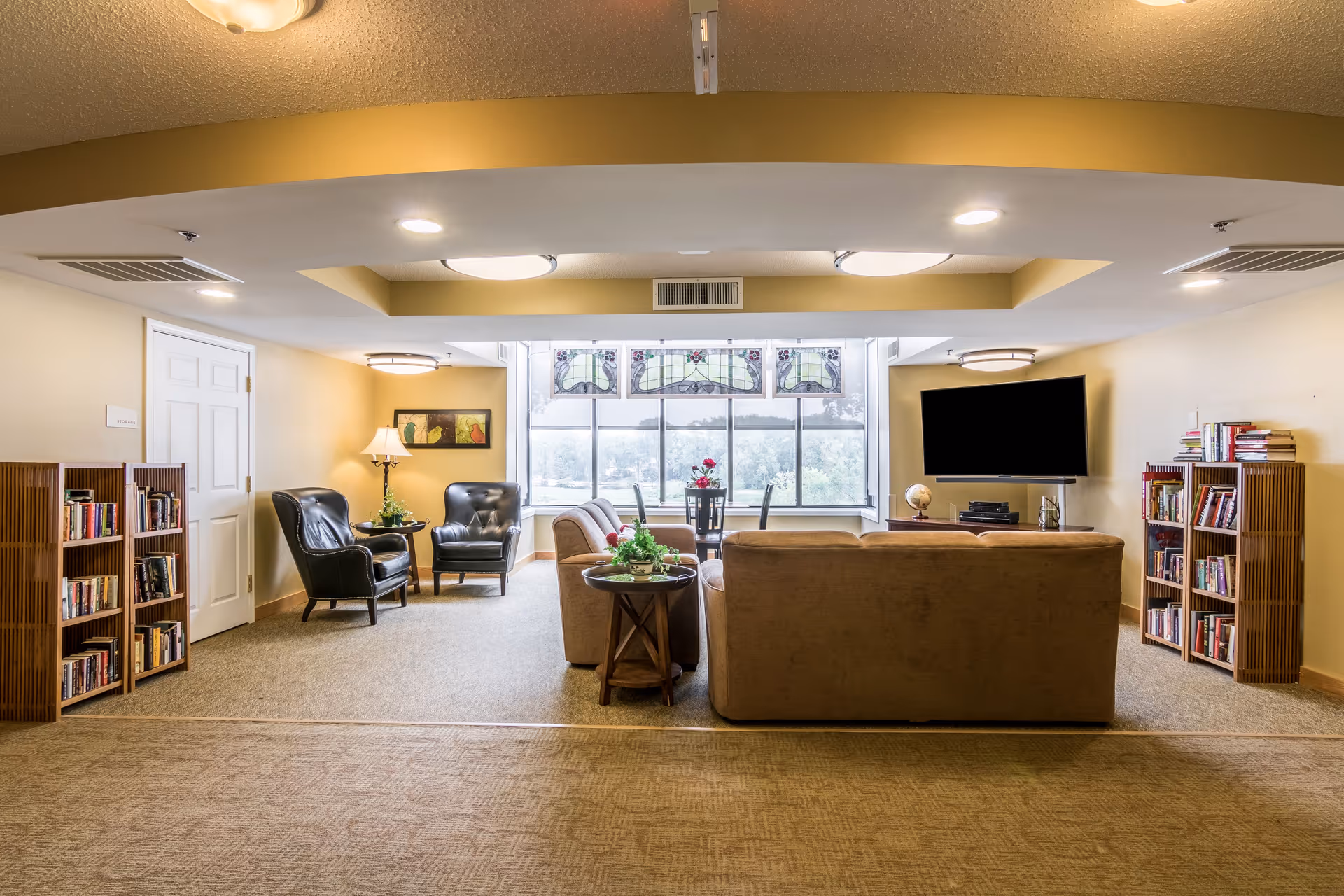 A cozy senior living common area with beige walls and carpet. The room features a large window with decorative stained glass panels at the top, letting in natural light. There are two black leather armchairs with a small table and lamp between them on the left side. In the center, a beige sofa faces a wall-mounted flat-screen TV. On either side of the TV, there are wooden bookshelves filled with books. A small round table with a plant is placed next to the sofa. The ceiling has recessed lighting and a tray ceiling design.