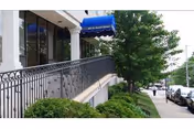 Exterior view of a building entrance with a blue awning, black metal railing along a ramp, green bushes, a tree, and parked cars along the street.