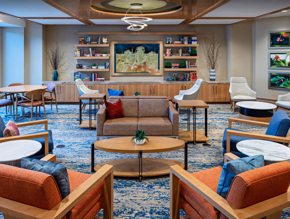 A spacious and well-lit living room area with modern furniture including sofas, armchairs, and small tables arranged on a blue patterned carpet. The back wall features wooden shelves filled with books and decorative items, a large framed artwork, and two tall vases with branches. The ceiling has a circular recessed design with a contemporary light fixture. On the right wall, there are three framed nature photographs.