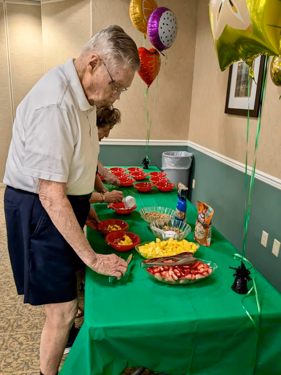 An elderly man and woman serving themselves fruit and cereal from bowls on a green tablecloth-covered table in a room decorated with colorful balloons.