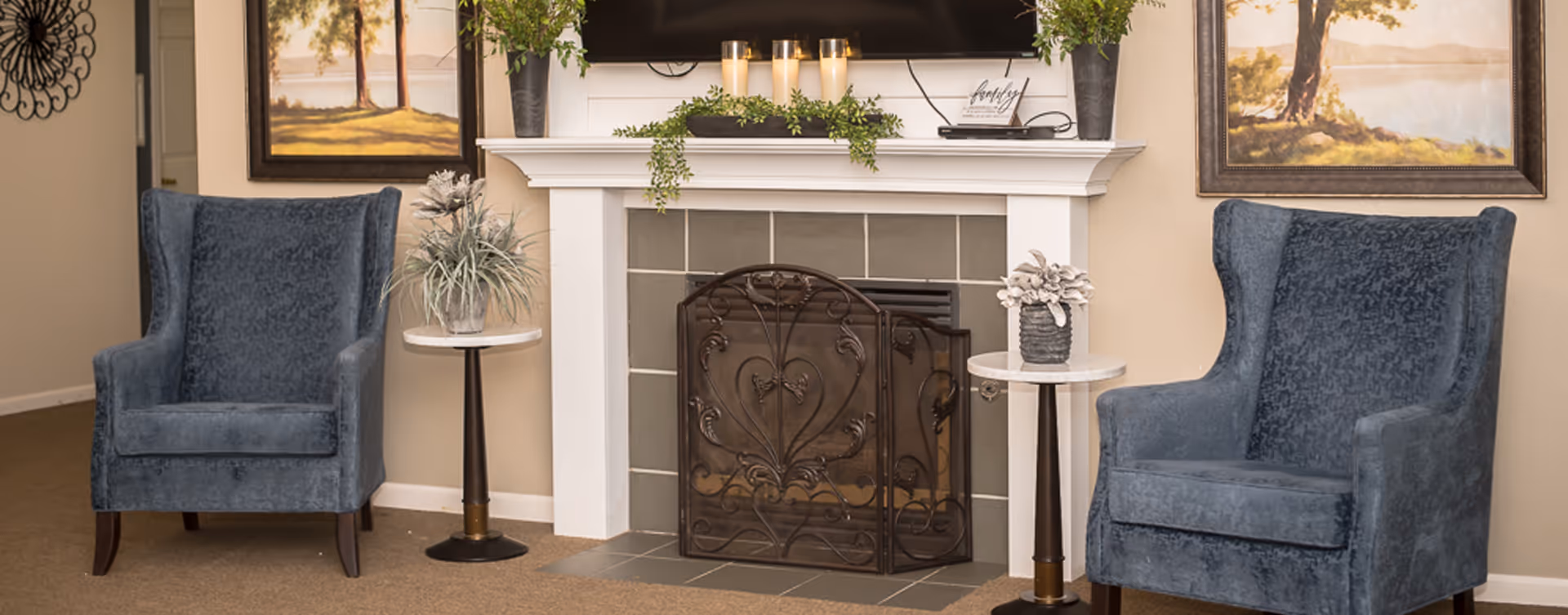 Two blue upholstered armchairs flank a decorative fireplace with a mantelshelf, plants, and small side tables.