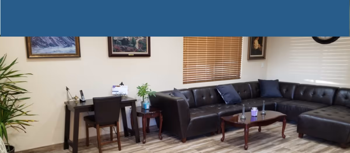 A living room with a large black leather sectional sofa adorned with dark blue pillows. In front of the sofa is a wooden coffee table with decorative items. To the left, there is a small desk with a chair and a potted plant on a side table. The walls are decorated with framed paintings and a window with wooden blinds is visible.