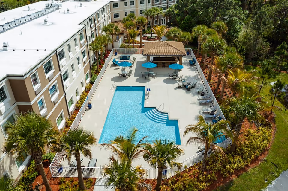 Aerial view of a rectangular outdoor swimming pool and surrounding patio with umbrellas, lounge chairs, palm trees, and a nearby multi-story building.